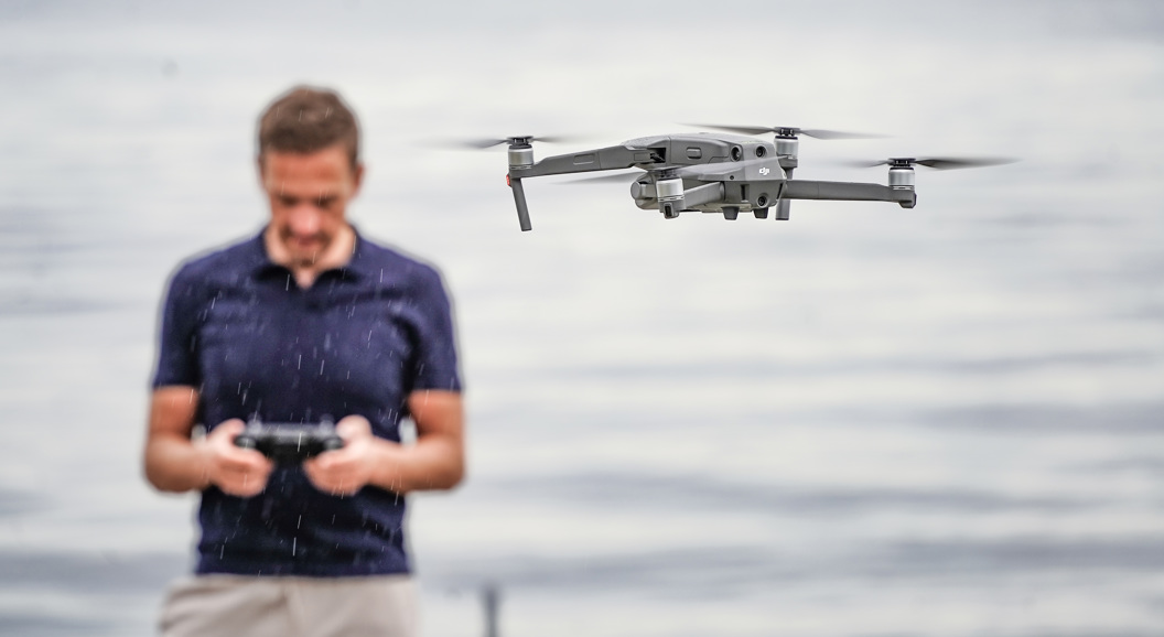 Kvinne i oransje jakke og strikket lue holder en drone i snødekte omgivelser.
