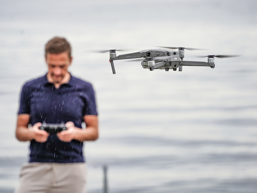 Kvinne i oransje jakke og strikket lue holder en drone i snødekte omgivelser.